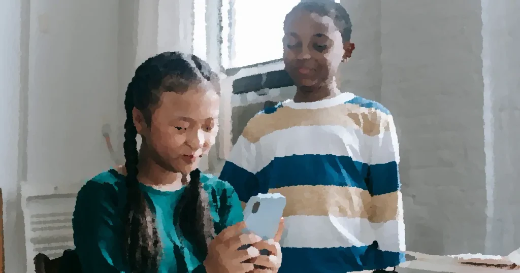 Happy little African American boy standing near Asian classmate girl using smartphone sitting at desk during break