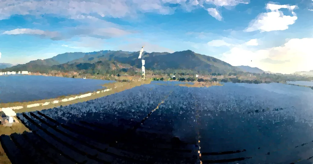 Aerial view of a vast solar farm with wind turbines against a mountainous backdrop on a sunny day.