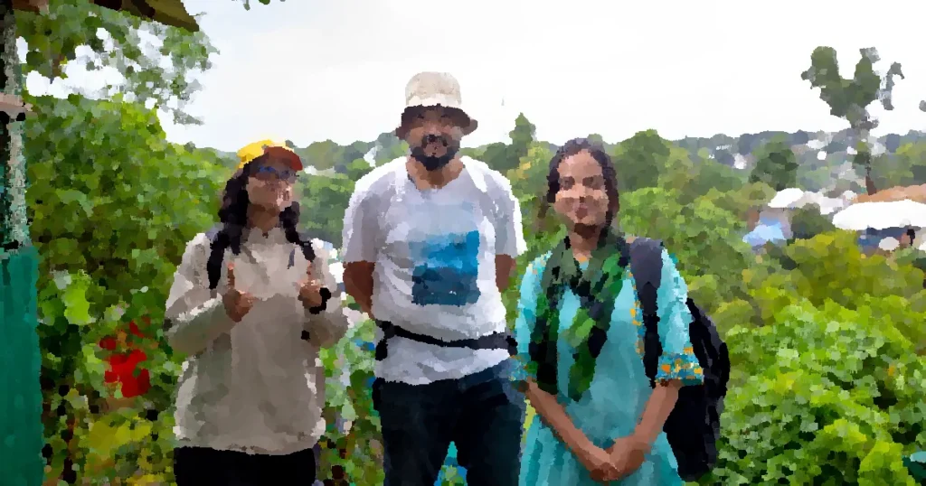 Three adults stand together smiling at a Bangladeshi refugee camp amidst lush greenery.